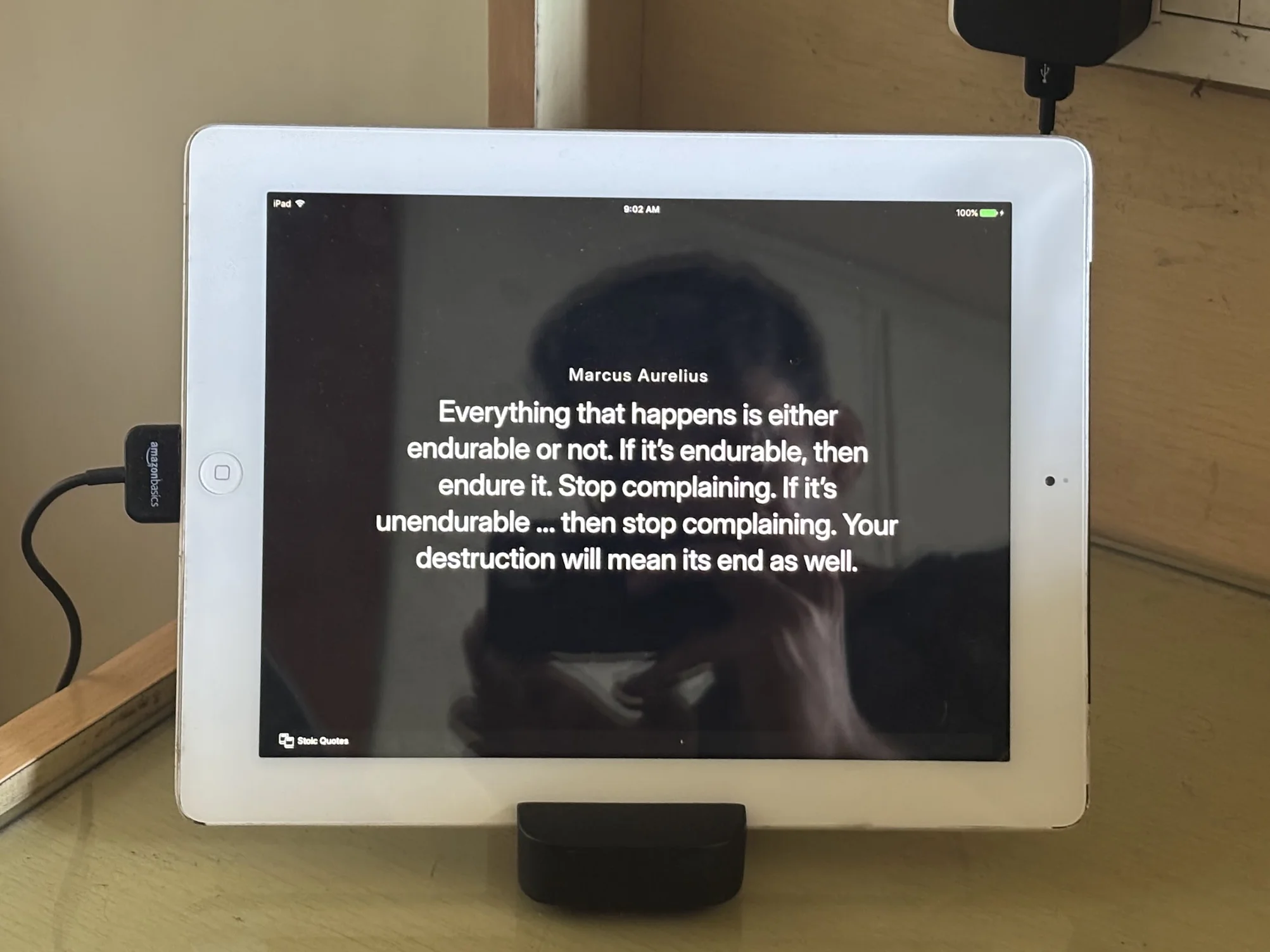 A white iPad sits on a wooden surface, connected to a charging cable, displaying a dark screen with a quote attributed to Marcus Aurelius. The quote is centered in white text, and a faint reflection of the person taking the photo is visible on the screen.